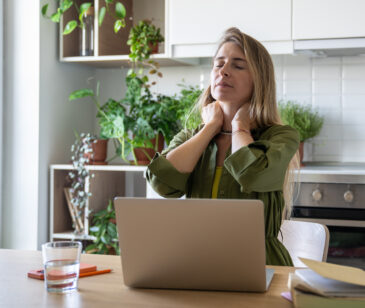 Frau steht mit Laptop und Wasserglas in der Küche und greift sich mit beiden Händen in den Nacken.