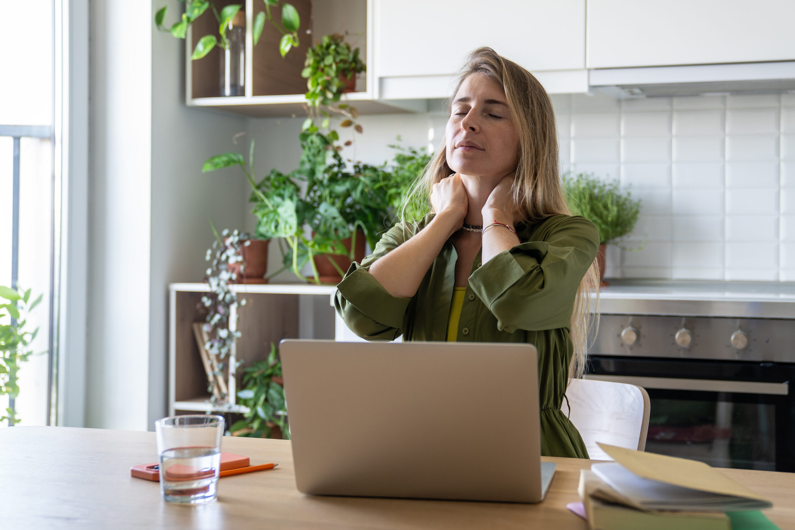 Frau steht mit Laptop und Wasserglas in der Küche und greift sich mit beiden Händen in den Nacken.