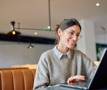 Frau mit Brille sitzt mit Laptop an einem Schreibtisch und lächelt.