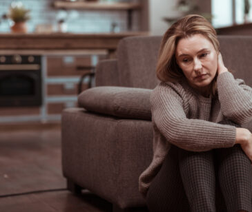 Frau sitzt neben dem Sofa mit der Hand im Nacken und schaut unglücklich zur Seite.