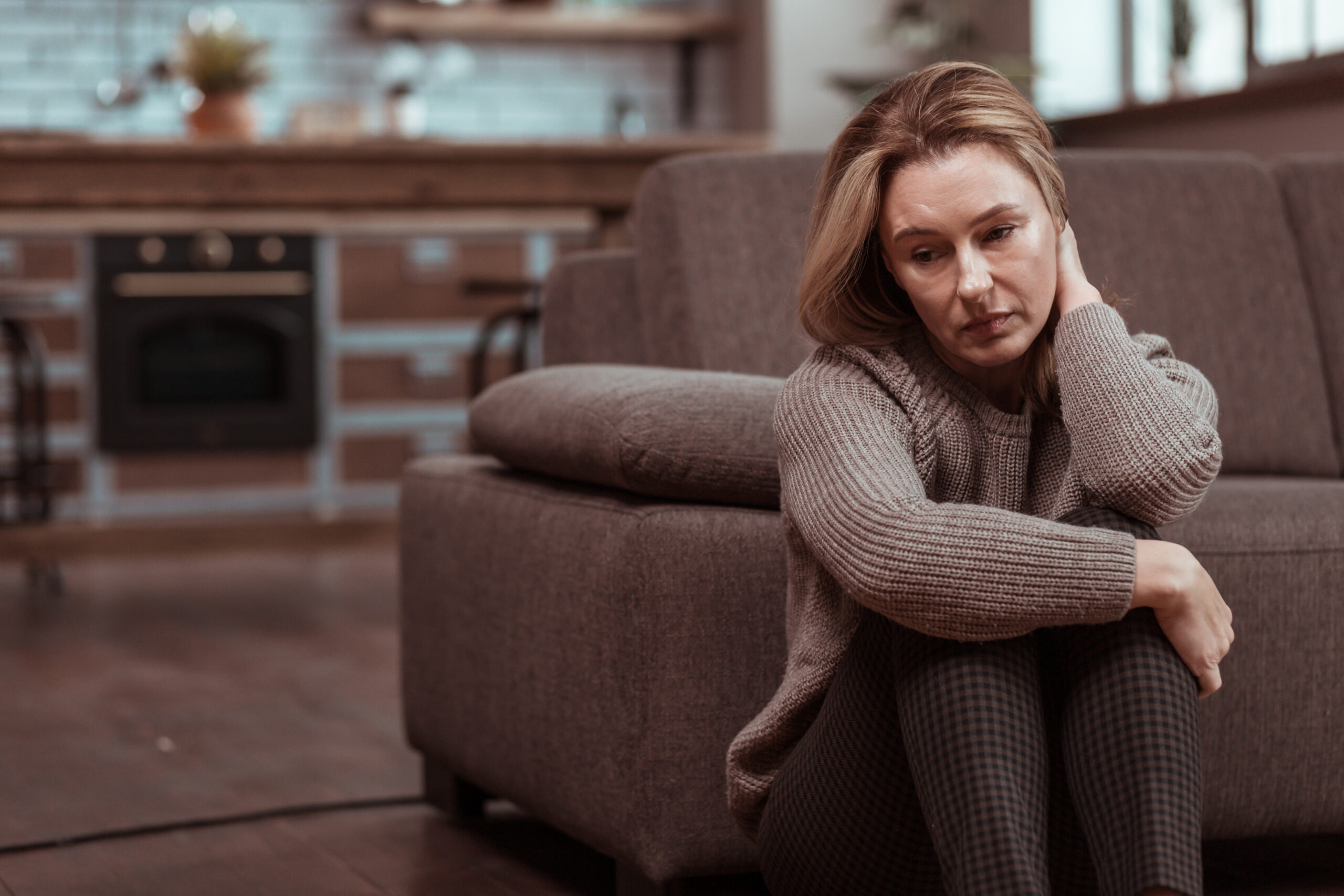Frau sitzt neben dem Sofa mit der Hand im Nacken und schaut unglücklich zur Seite.