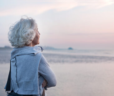 Eine Frau steht am Strand mit dem Blick auf das Meer gerichtet. Es ist nur Das Profil der Frau zu sehen.