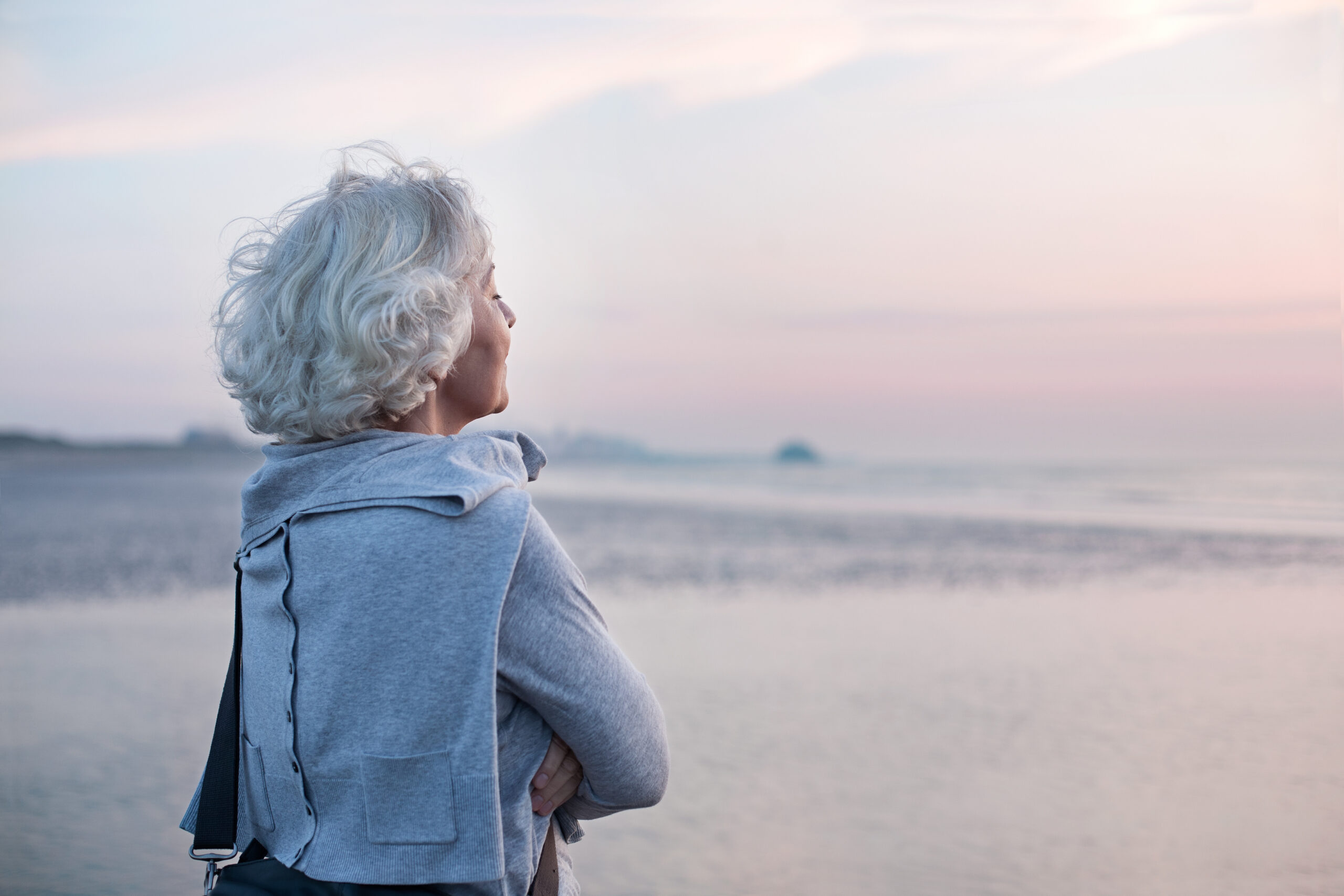 Eine Frau steht am Strand mit dem Blick auf das Meer gerichtet. Es ist nur Das Profil der Frau zu sehen.
