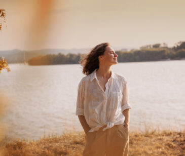 Frau mittleren Alters steht in einer weiten Bluse an einem See und schaut mit geschlossenen Augen genussvoll nach rechts mit.