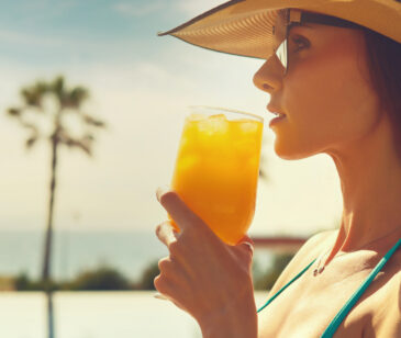 Frau mit Sonnenbrille und Hut trinkt aus einem Cocktailglas mit Eiswürfeln und orangener Flüssigkeit.