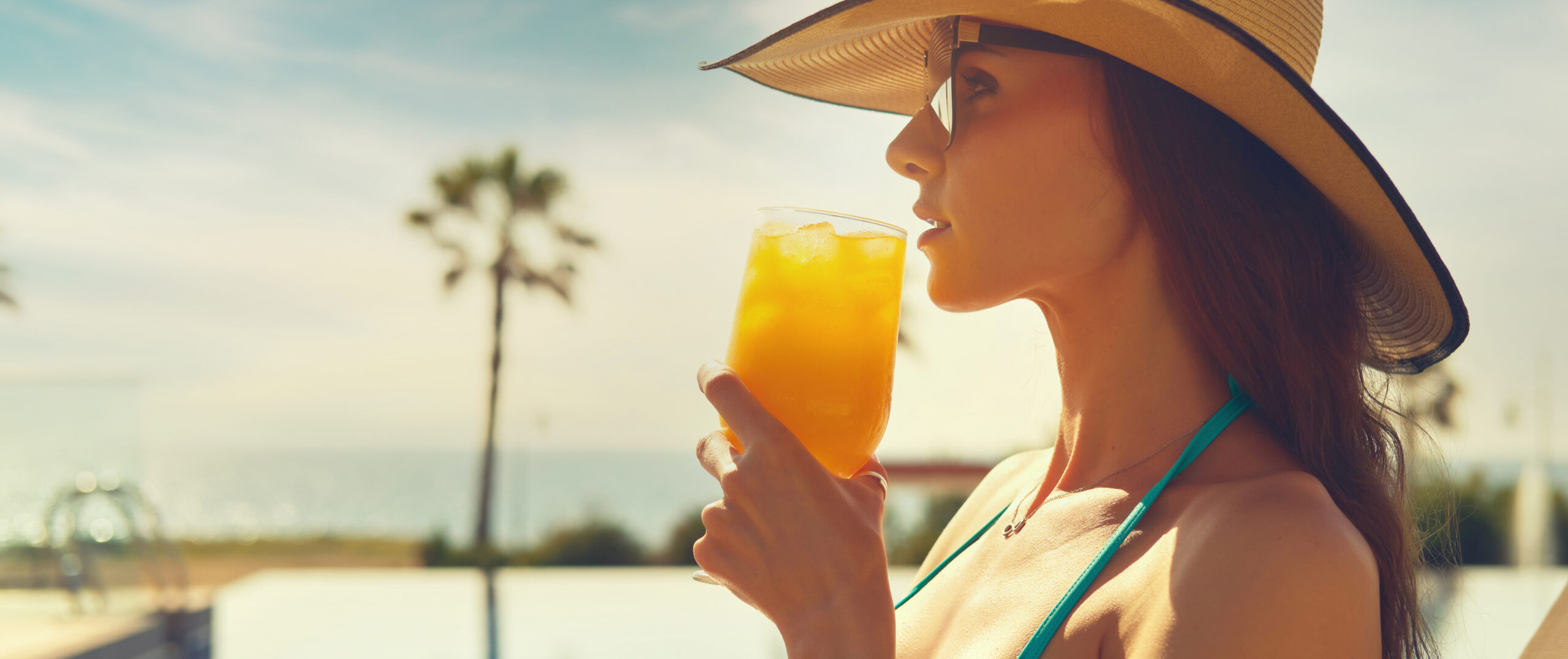 Frau mit Sonnenbrille und Hut trinkt aus einem Cocktailglas mit Eiswürfeln und orangener Flüssigkeit.