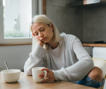 Junge Frau mit Eisenmangel sitzt am Esstisch, mit einer Tasse in der Hand, sieht müde und blass aus.
