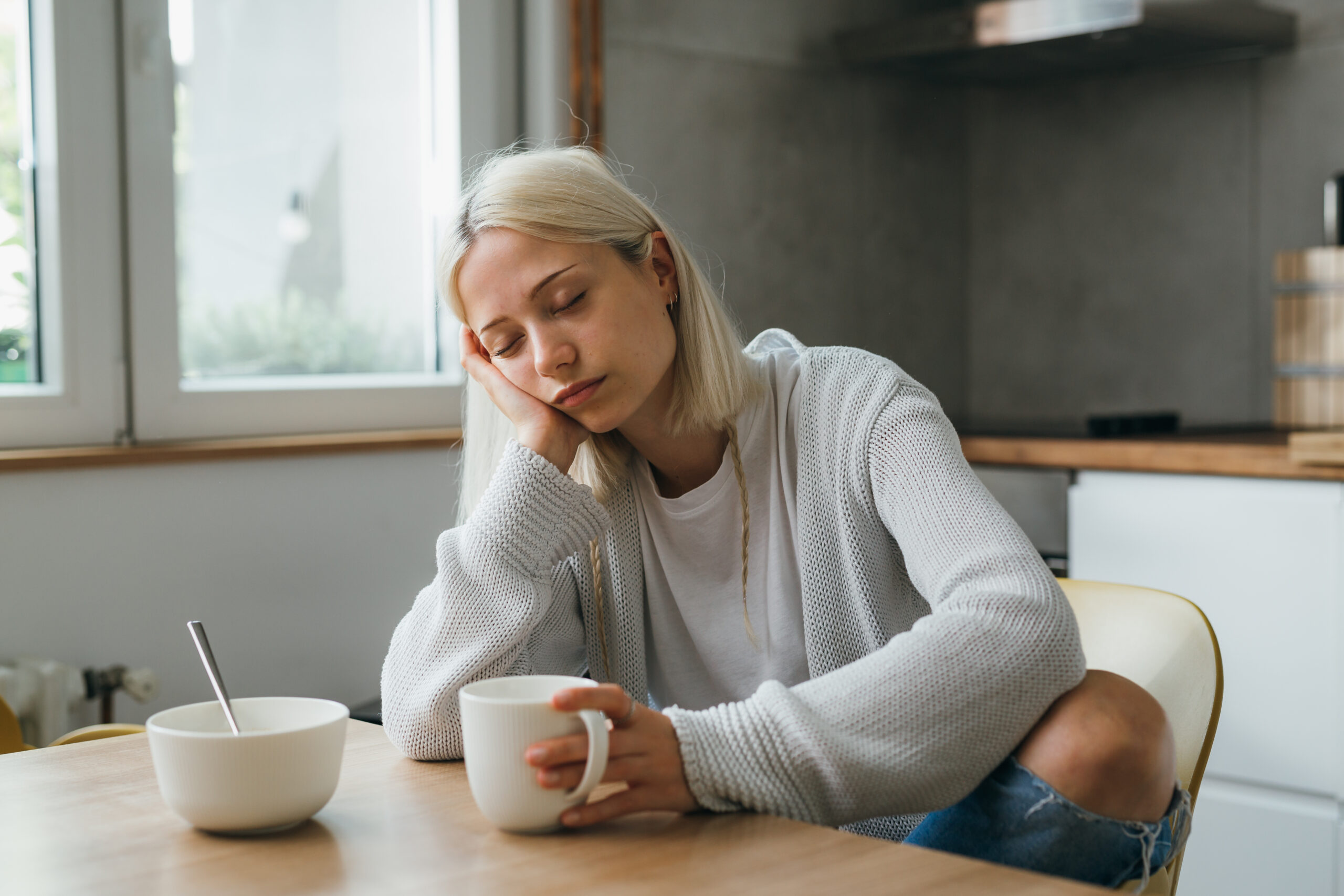 Junge Frau mit Eisenmangel sitzt am Esstisch, mit einer Tasse in der Hand, sieht müde und blass aus.