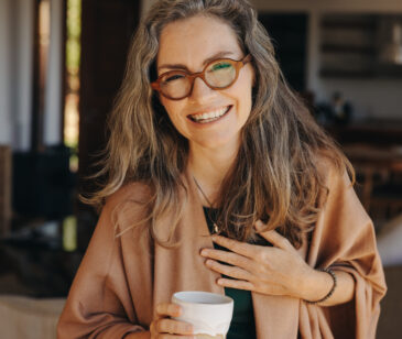Frau mittleren Alters mit brauner Brille und langen braunen Haaren lächelt glücklich in die Kamera. Sie hält eine Tasse in der Hand und legt die Hand auf ihre Brust.