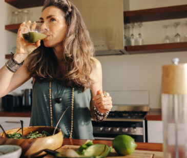 Frau mittlereln Alters steht in der Küche und trinkt einen grünen frisch gepressten Saft.